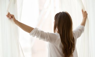 Rear view at woman opening window curtains at home or hotel starting new day, enjoying wellbeing or light good pleasant morning looking out feeling happy, breathing fresh air, relaxing on weekend