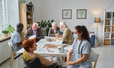Nurse talking to elderly woman during medical exam with other senior people in the background in nursing home